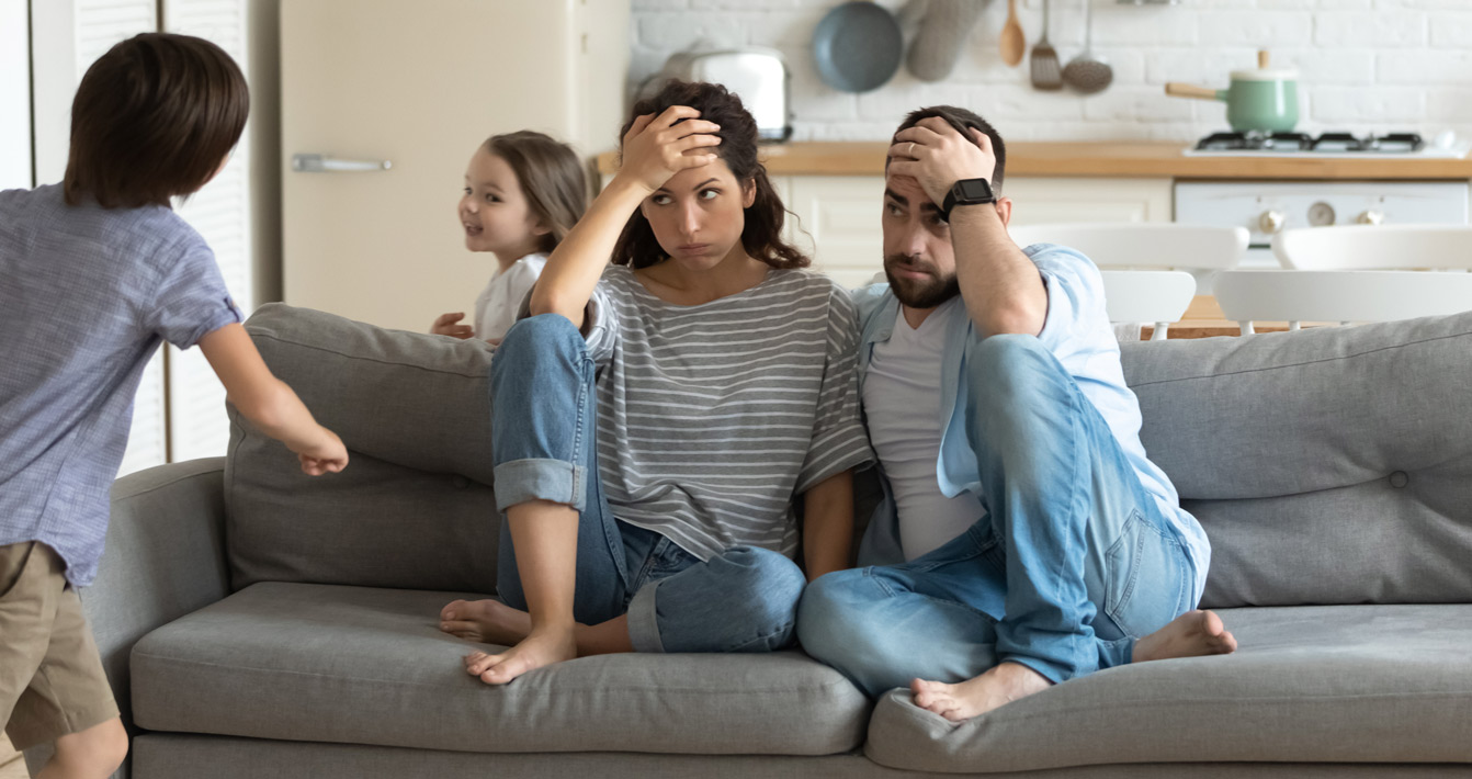 Gestresste junge Eltern, die auf der Couch saßen, müde von verrückten lauten hyperaktiven kleinen Kindern, Geschwistern zu Hause. Unglückliches Ehepaar, irritiert von ungezogenem Nachwuchs, die an Kopfschmerzen leidet.