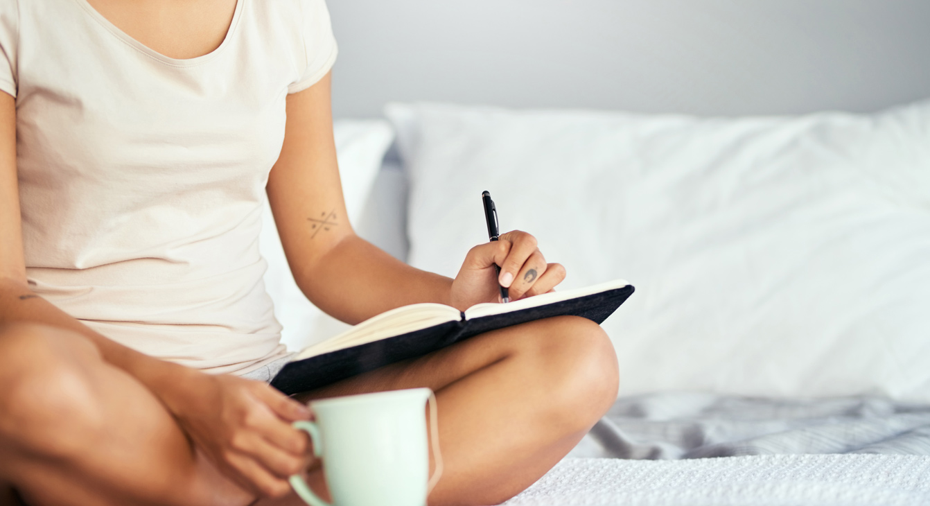 Ausschnitt einer linkshändigen Frau, die morgens auf ihrem Bett sitzt und in ein Tagebuch schreibt.