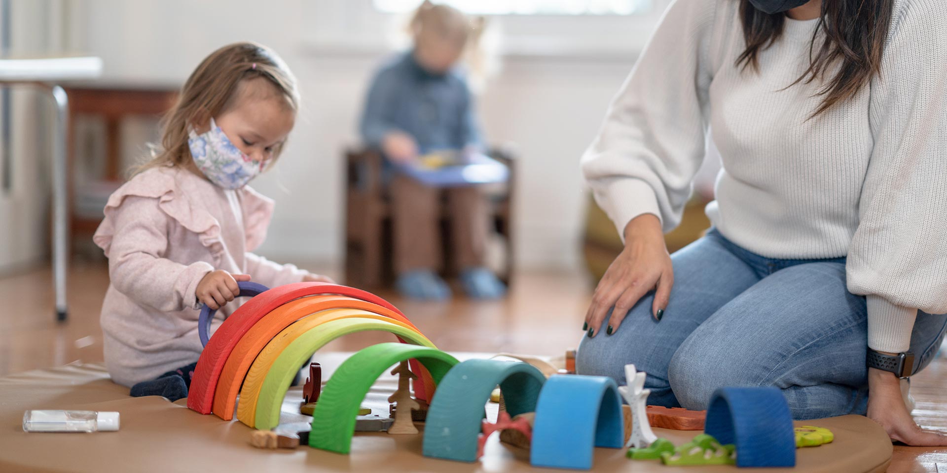 a preschool teacher is playing with an arch rainbow tunnel on the floor with a preschool girl. they are both wearing a face mask during the coronavirus pandemic to prevent the spread of germs.
