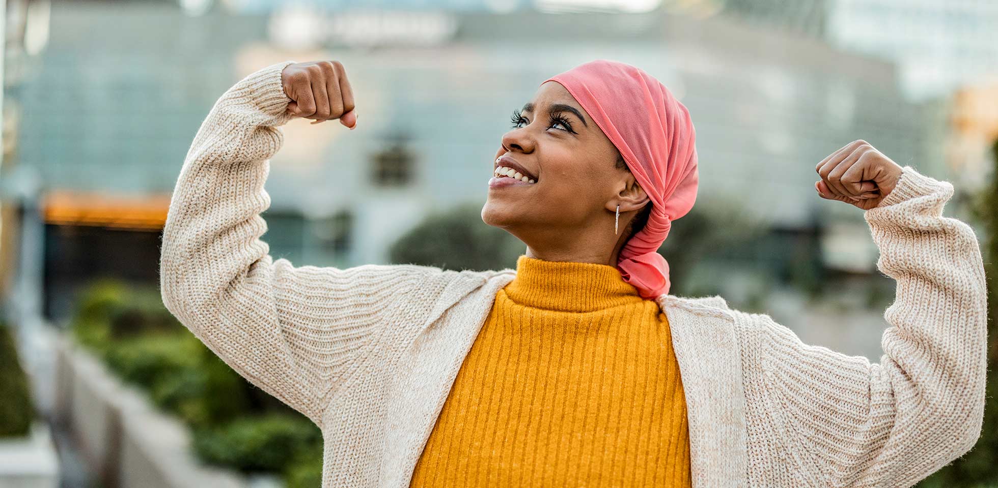 une survivante du cancer joyeuse, âgée d'une trentaine d'années, portant un foulard rose et un pull jaune, qui montre ses muscles dans un décor urbain.