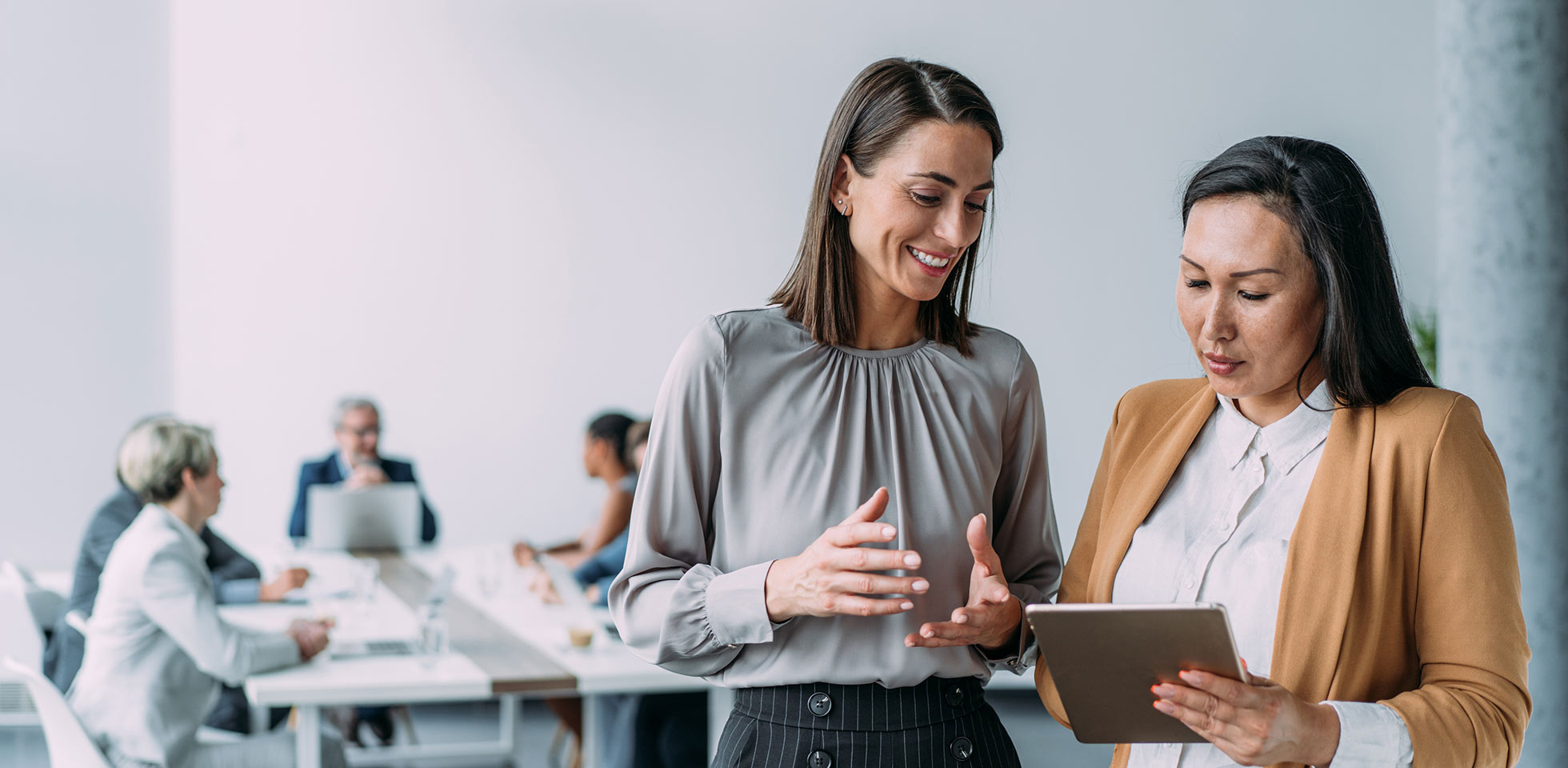 photo de deux collègues féminines en pleine discussion dans un bureau mone. femmes d'affaires en réunion utilisant une tablette numérique et discutant de stratégie commerciale. professionnels confiants travaillant ensemble au bureau. cadres d'entreprise discutant d'un nouveau projet et échangeant des idées sur leur lieu de travail. femmes d'affaires accomplies debout avec leurs collègues travaillant en arrière-plan.
