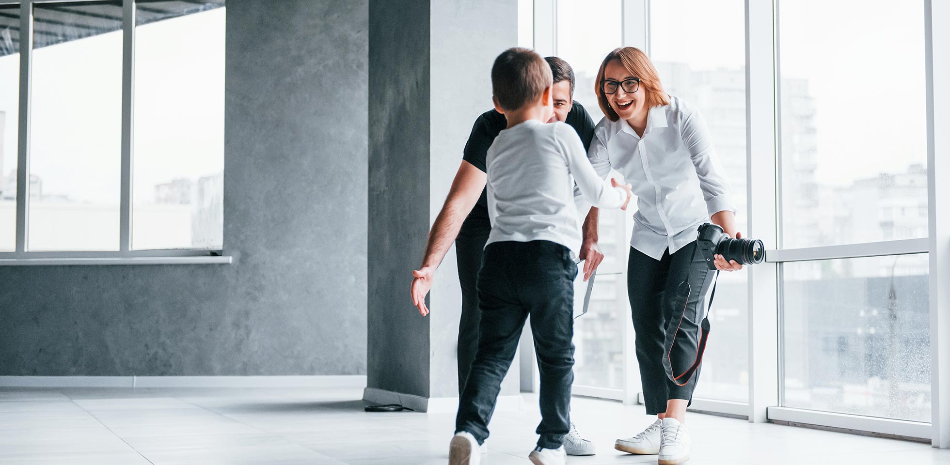 femme en tenue formelle, appareil photo à la main, debout dans une pièce vide avec un homme et un petit garçon qui courent et s'amusent.
